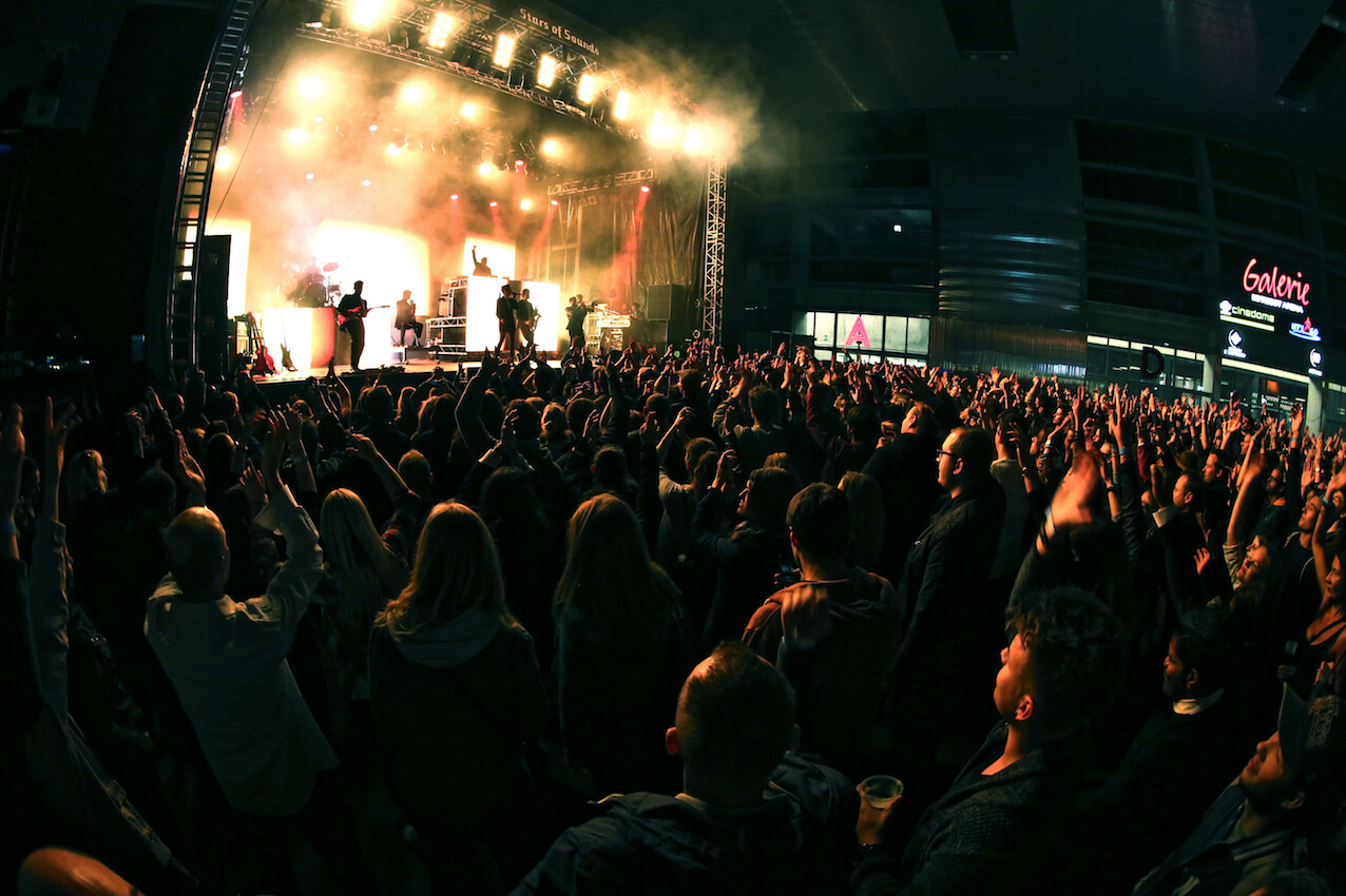 Eine große Menschenmenge steht bei einem Hallenkonzert vor einer hell erleuchteten Bühne, auf der Künstler auftreten und Menschen ihre Hände in die Luft strecken.