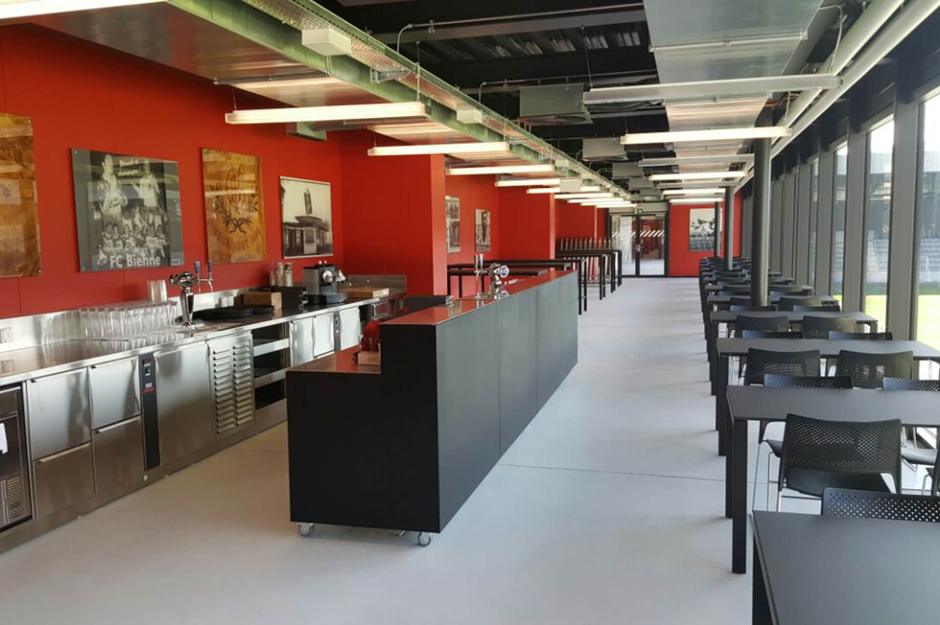 Moderne Cafeteria mit roten Wänden, schwarzen Tischen und Stühlen, großen Fenstern auf einer Seite und einer langen zentralen Theke mit Küchengeräten aus Edelstahl.