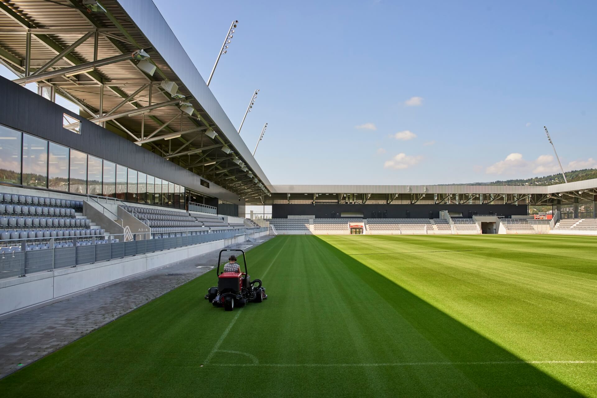 Eine Person bedient einen Rasenmäher auf dem Rasen eines leeren, modernen Stadions unter freiem Himmel.