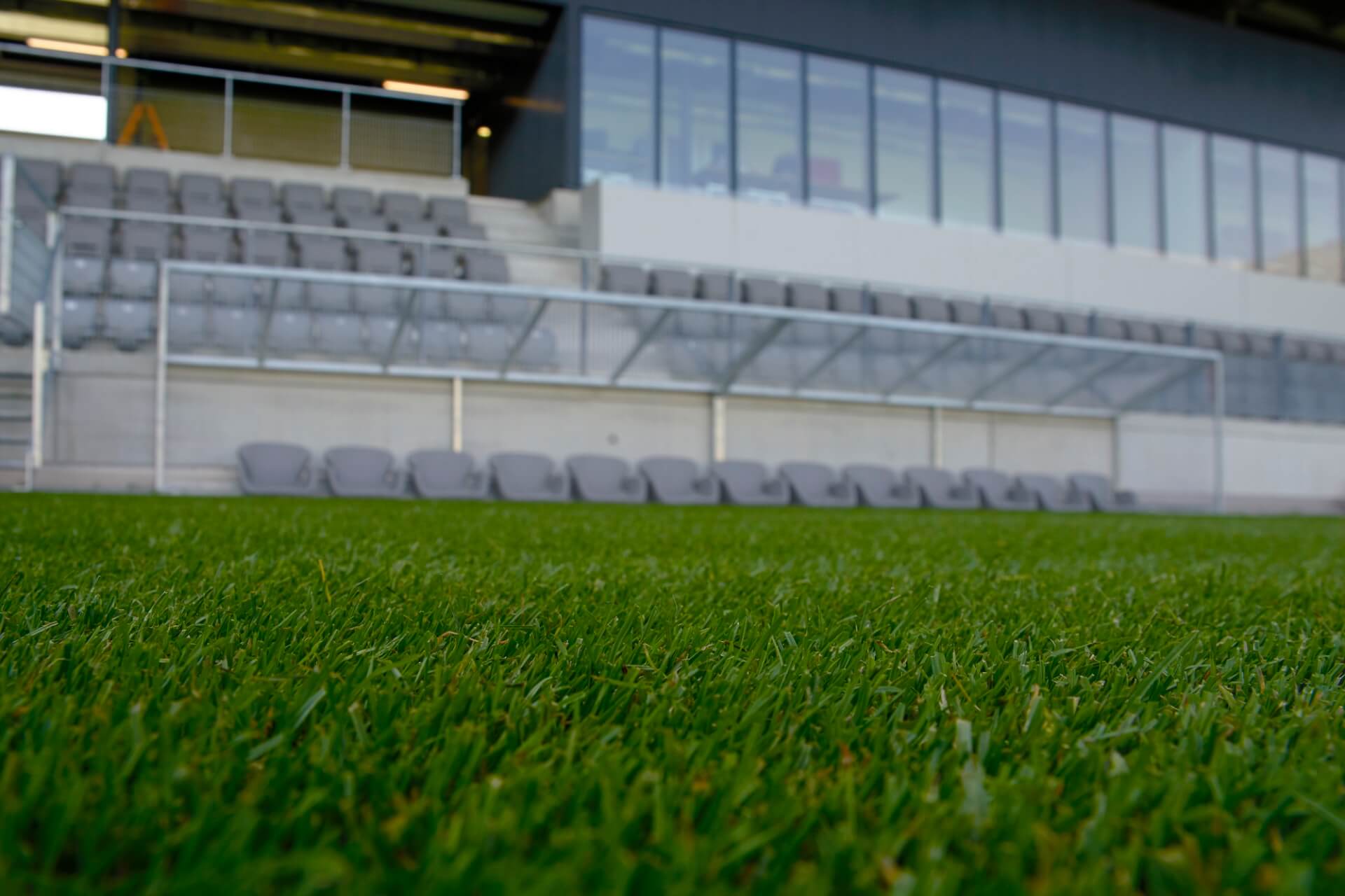 Leere Stadionbänke und Sitzplätze am Rande eines grünen Fußballfelds, mit grauen Sitzen und einem Glasdach im Hintergrund.