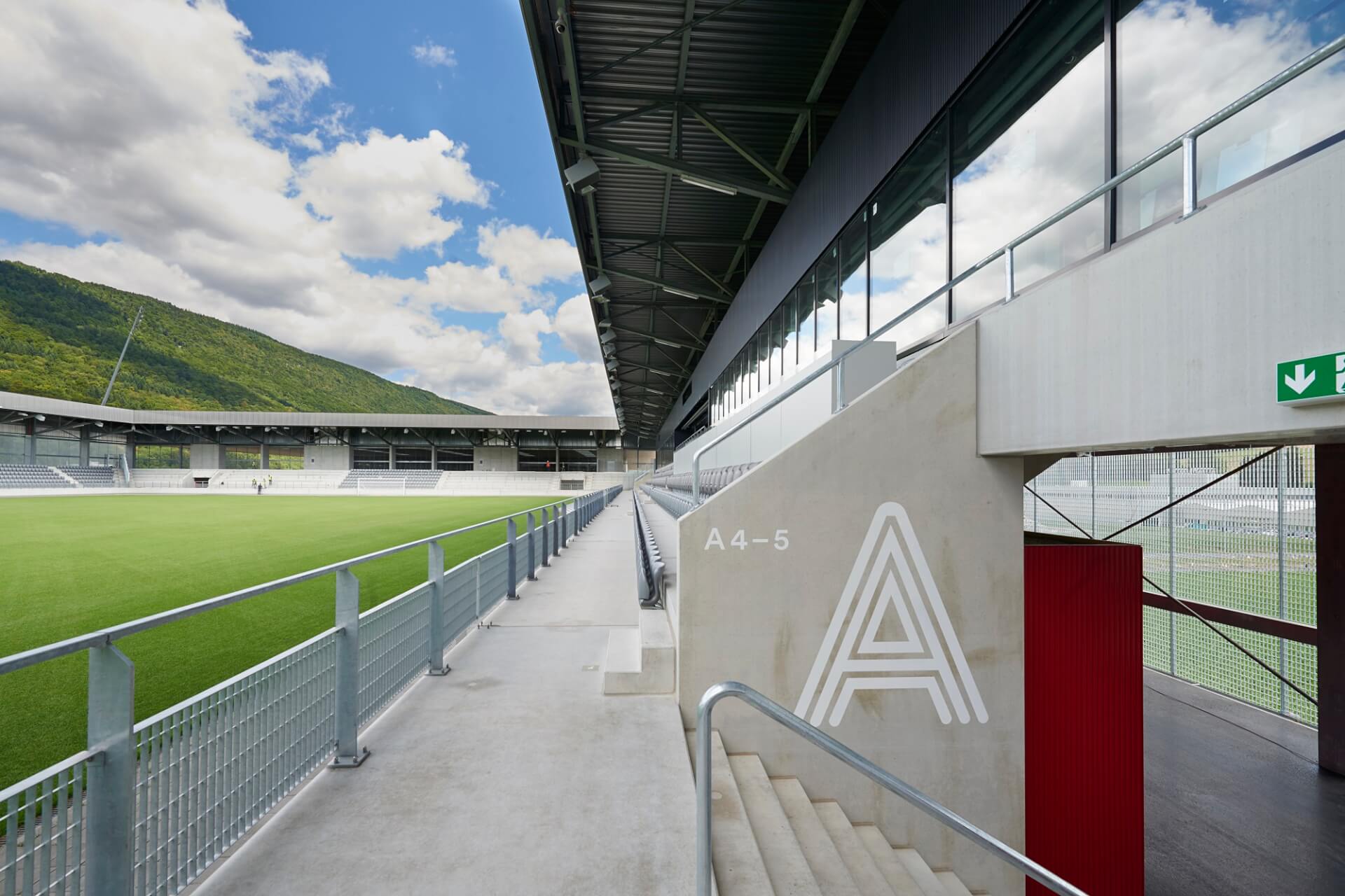 Ein modernes Sportstadion im Freien mit grünem Rasen, überdachten Sitzplätzen, Betonstegen und klarer Beschilderung. Im Hintergrund sind Berge und Wolken zu sehen.