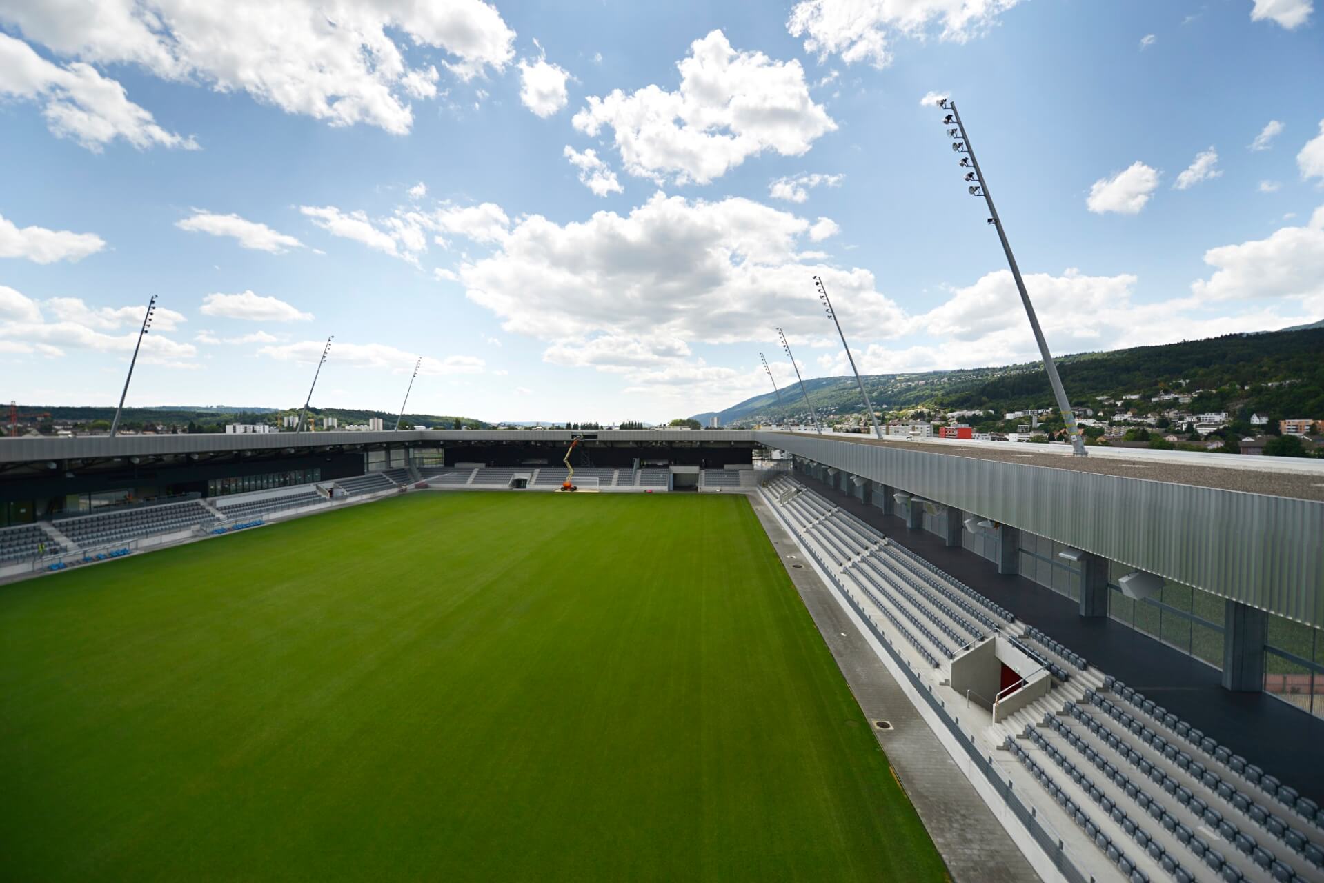 Ein leeres Fußballstadion mit grünem Rasen, grauen Sitztribünen, hohen Flutlichtern und einem teilweise bewölkten Himmel im Hintergrund.