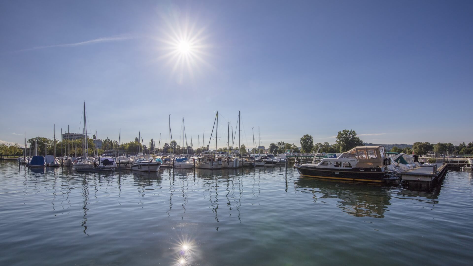 Ein Yachthafen mit mehreren angedockten Booten bei strahlend blauem Himmel und heller Sonne, die sich auf dem ruhigen Wasser spiegelt.