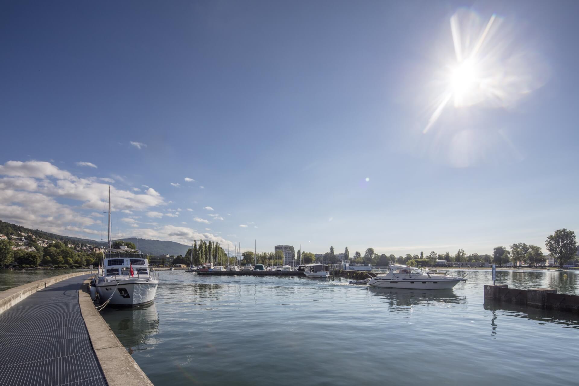 Ein kleiner Jachthafen mit mehreren angedockten Booten bei strahlend blauem Himmel und heller Sonne, mit ruhigem Wasser und einer entfernten Uferlinie mit Bäumen und Gebäuden.