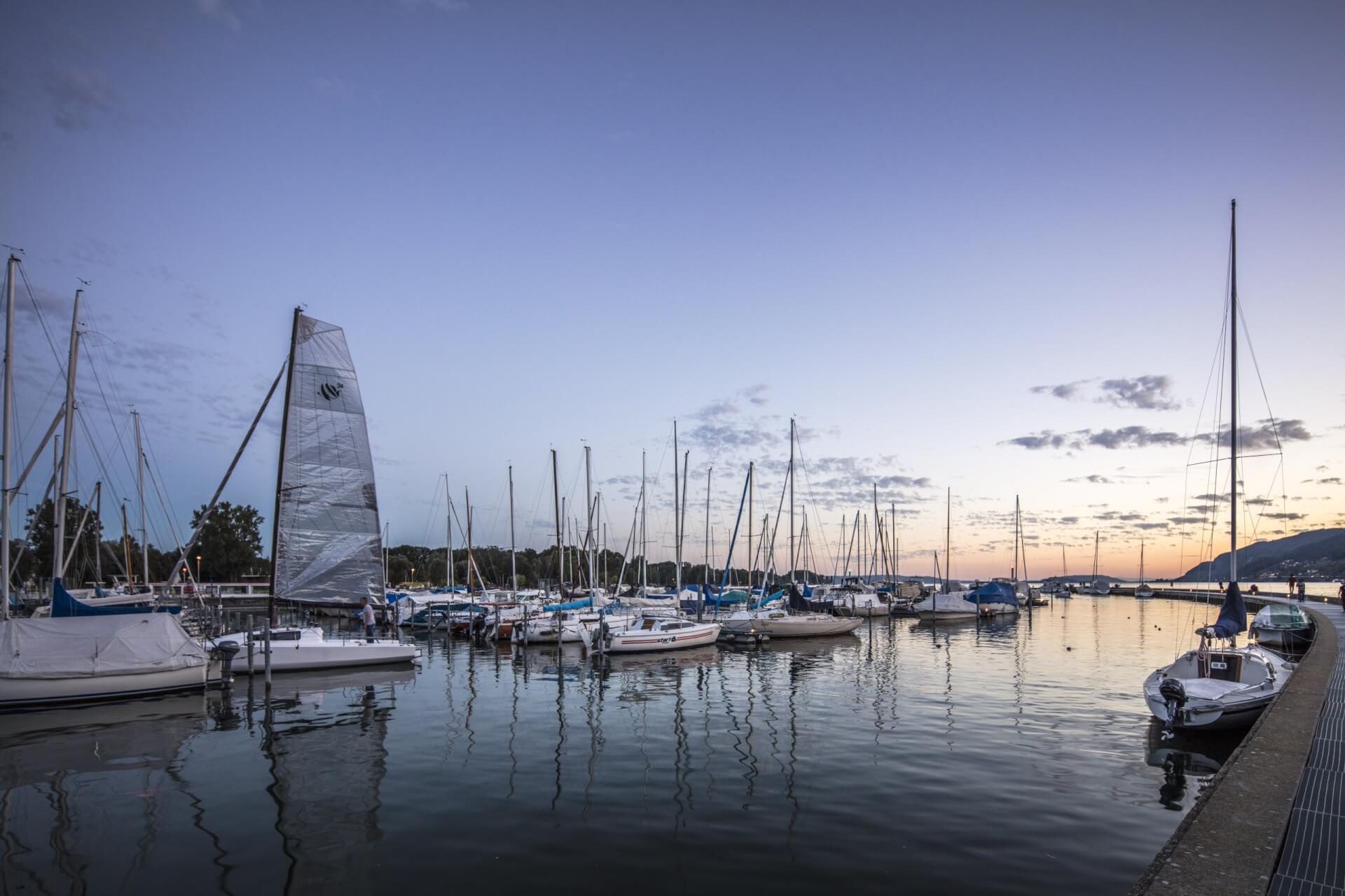 Segelboote und Yachten in einem Yachthafen bei Sonnenuntergang, mit ruhigem Wasser, in dem sich die Boote spiegeln, und einem klaren Abendhimmel.