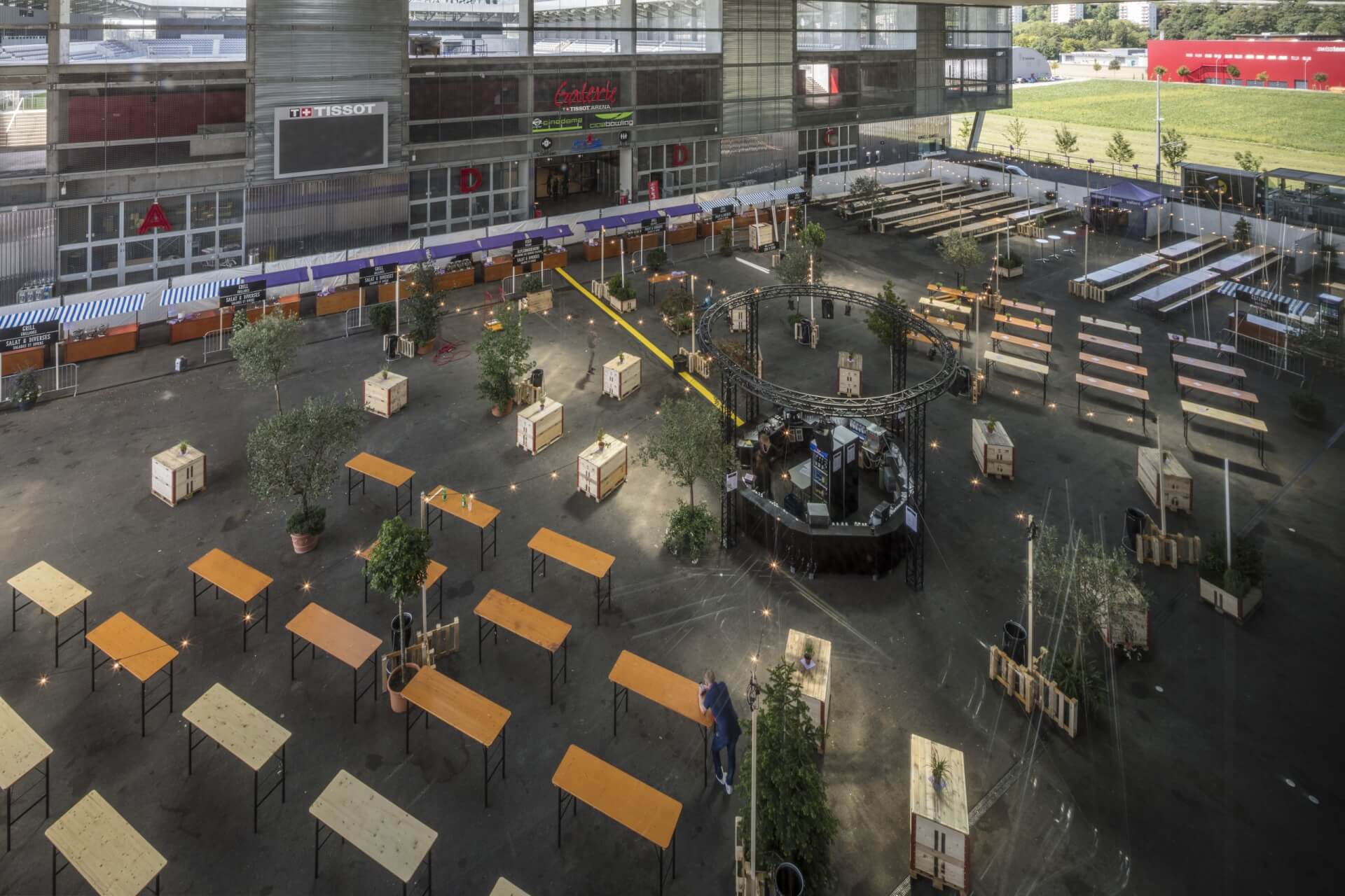 Großer Innenraum mit vielen leeren Holztischen und -bänken, einer kleinen runden Bühne mit Musikanlage und einigen getopften Bäumen; es ist kein Publikum anwesend.