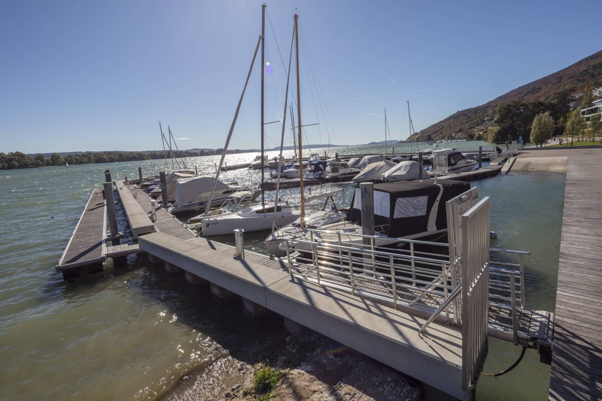 Ein kleiner Jachthafen mit mehreren angedockten Booten und Segelbooten an einem sonnigen Tag, mit Hügeln im Hintergrund und ruhigem Wasser im Vordergrund.