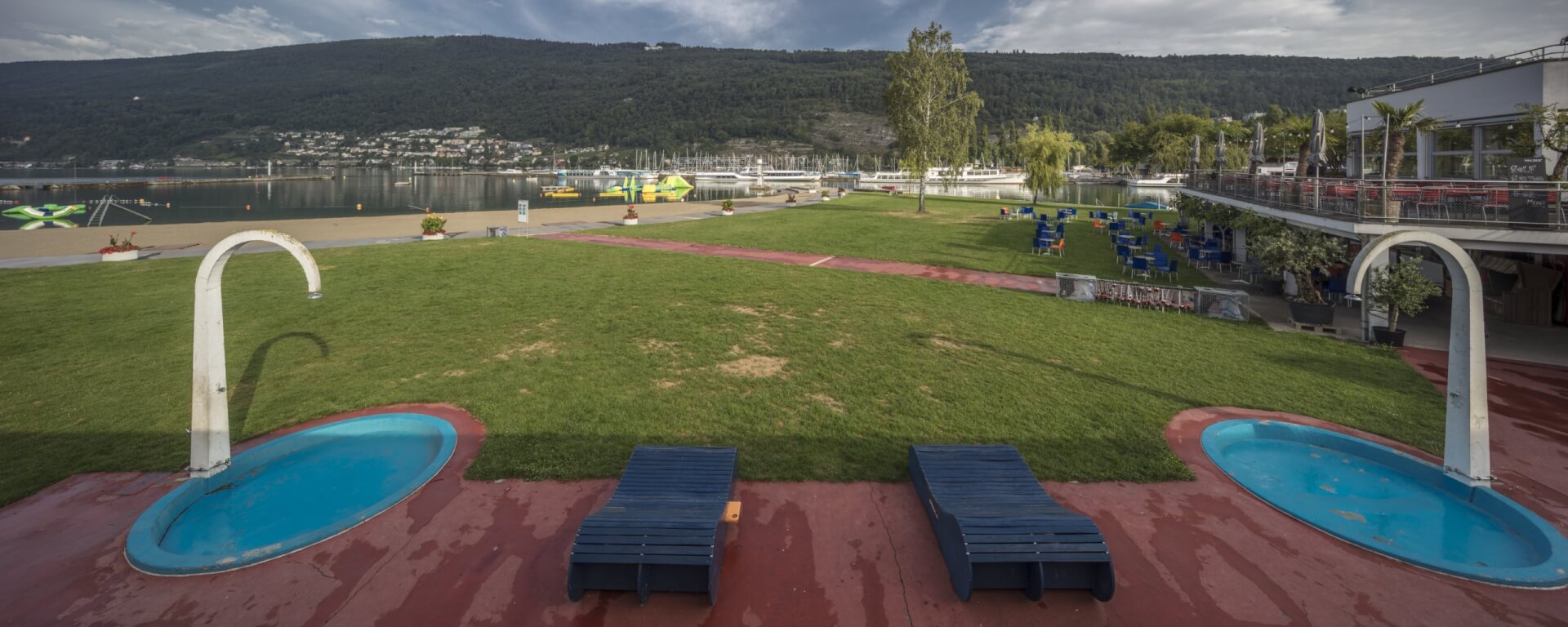 Deux chaises longues bleues sont placées entre deux petites piscines circulaires sur un patio rouge, donnant sur une zone herbeuse, une plage et un lac avec des bateaux et des collines en arrière-plan.