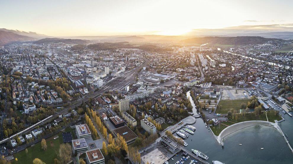 Luftaufnahme einer Stadt bei Sonnenuntergang mit dicht gedrängten Gebäuden, einem Fluss mit Booten und einer Parkanlage in der Nähe des Wassers. Im Hintergrund sind die Berge zu sehen.