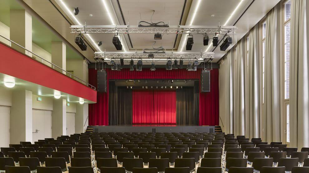 Ein modernes Auditorium mit Reihen leerer schwarzer Stühle vor einer Bühne mit roten Vorhängen, großen Fenstern auf einer Seite und Oberlicht.