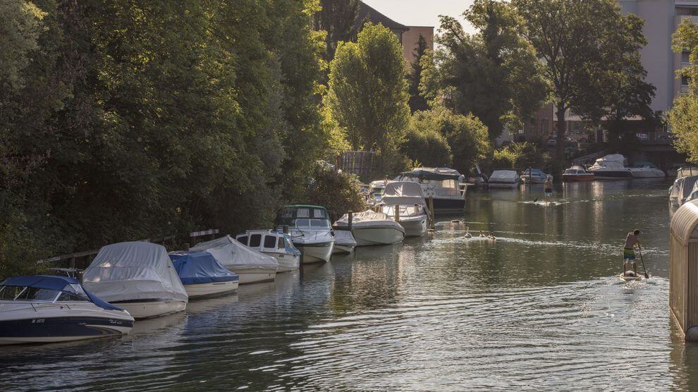 An einem von Bäumen gesäumten Kanal sind mehrere Boote vertäut, auf denen eine Person mit dem Paddleboard fährt.