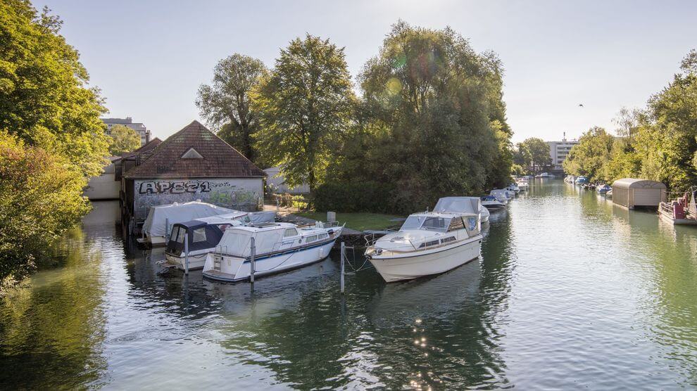 Mehrere Boote liegen an einem ruhigen Fluss, umgeben von Bäumen und einem Gebäude mit Graffiti, unter einem klaren Himmel.