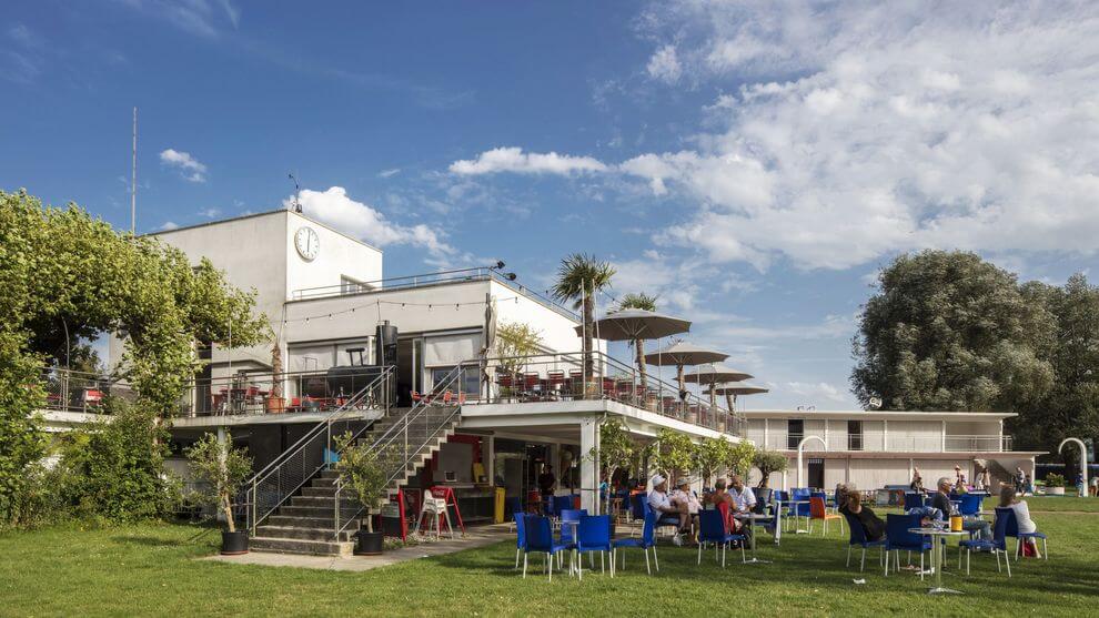 Bâtiment moderne de deux étages avec des places assises à l'extérieur sur les deux niveaux, les gens dînant sur une pelouse et un patio, sous un ciel partiellement nuageux.