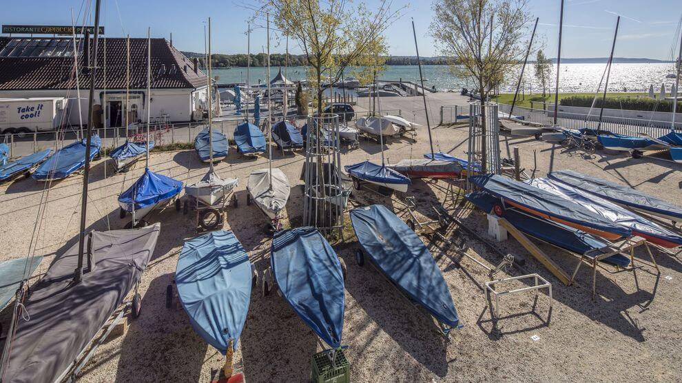 Eine Reihe kleiner, mit Planen abgedeckter Segelboote auf einem Kiesplatz am Seeufer; im Hintergrund Bäume und ein Gebäude mit einem 