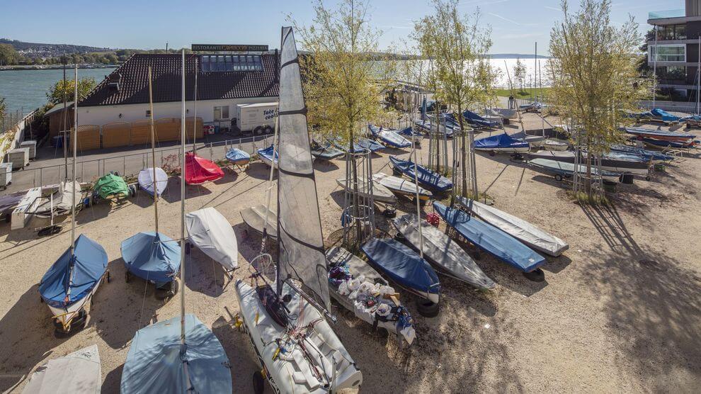 Eine Vielzahl kleiner Segelboote und Jollen, viele davon überdacht, sind auf einem sandigen Grundstück in der Nähe eines Flusses geparkt, mit Bäumen und Gebäuden in der Nähe und unter klarem Himmel.