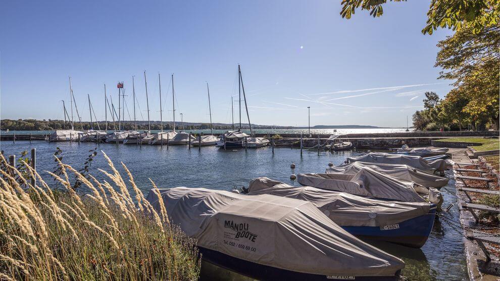 Ein kleiner Jachthafen mit überdachten Motorbooten im Vordergrund und angedockten Segelbooten im Hintergrund unter einem klaren blauen Himmel.
