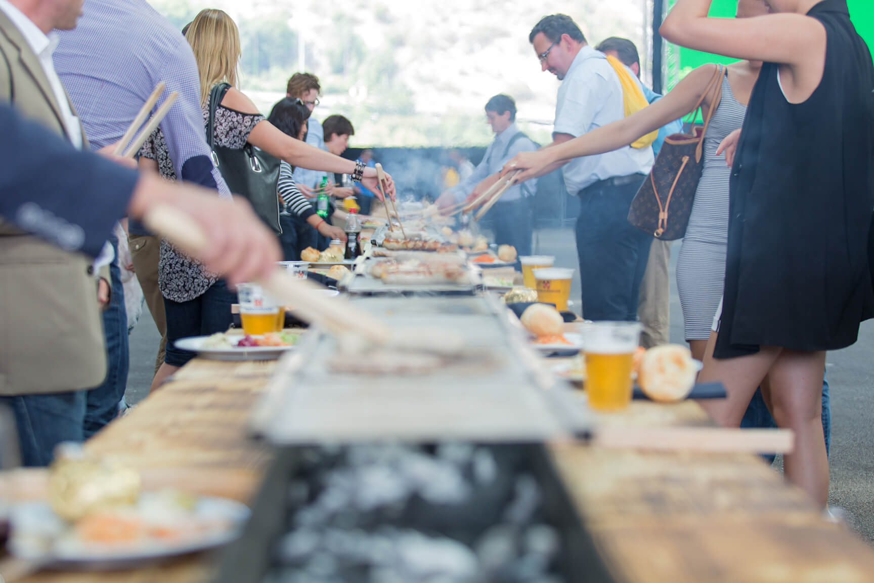Die Leute stehen um einen langen Grill im Freien und bereiten das Essen zu, auf den Holztischen daneben stehen Teller und Getränke.
