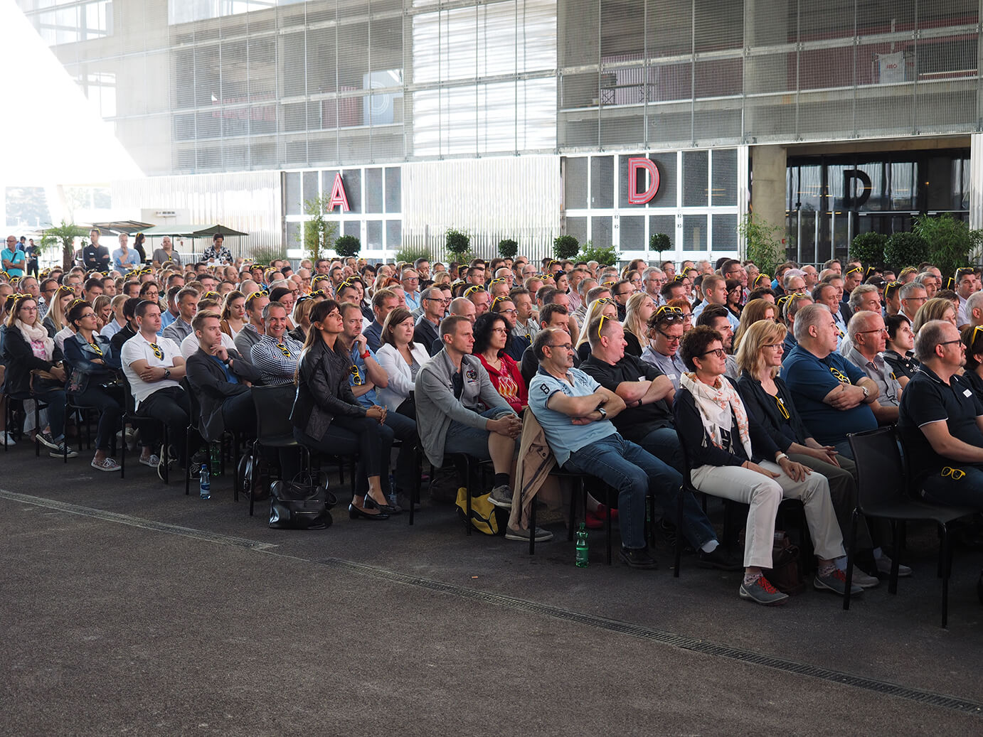 Ein großes Publikum sitzt dicht gedrängt in Stuhlreihen in einer geräumigen Halle mit Wänden im Industriestil und großen Fenstern.