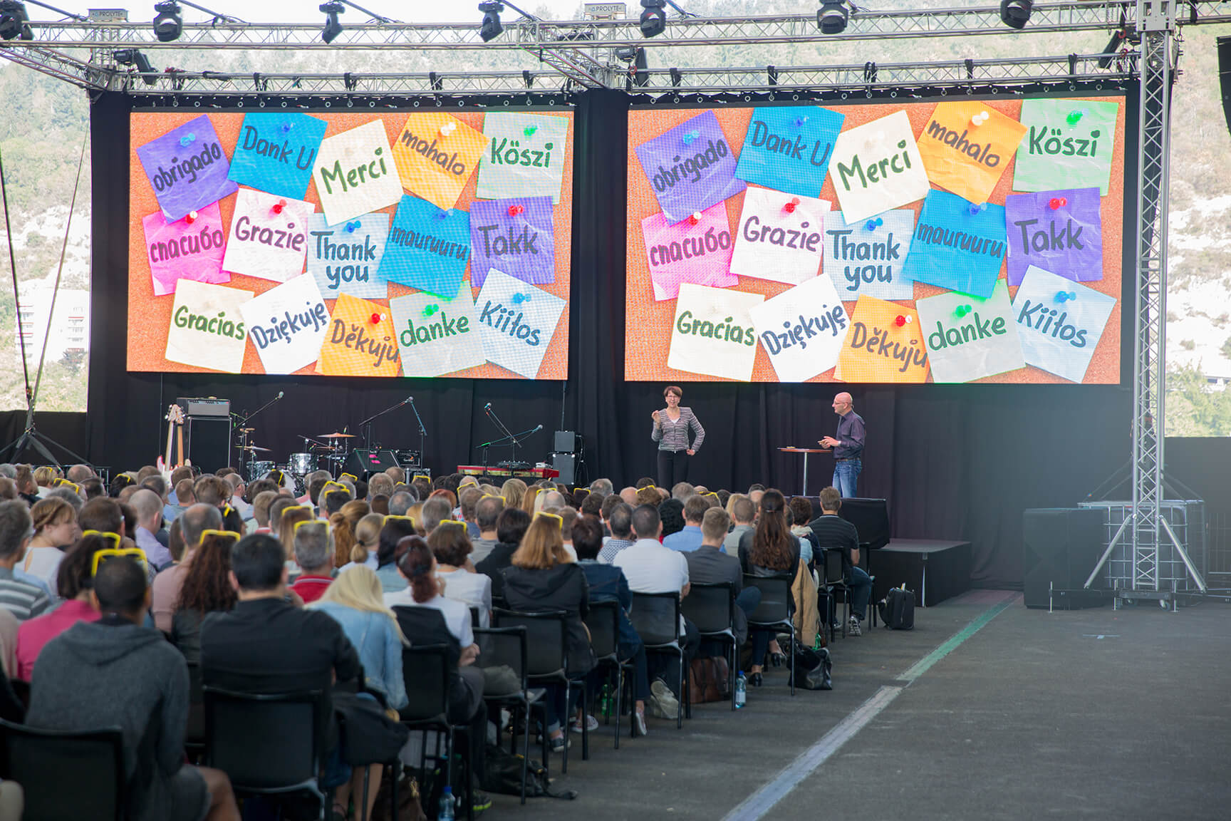 Ein Redner wendet sich an ein sitzendes Publikum im Freien, während hinter ihm zwei große Bildschirme auf bunten Karten Danksagungen in mehreren Sprachen anzeigen.