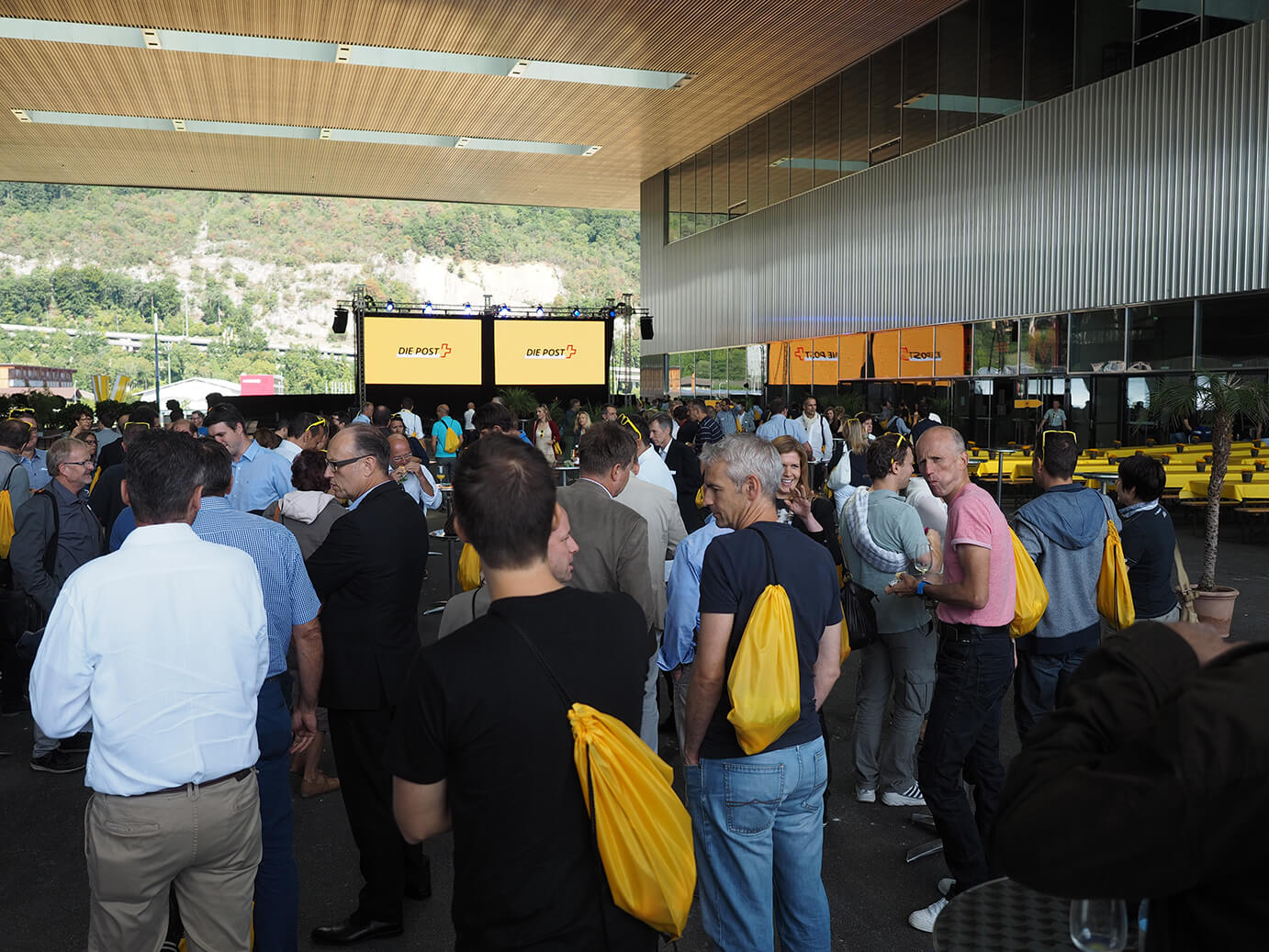 Eine große Gruppe von Menschen versammelt sich auf einer Veranstaltung im Freien, viele tragen gelbe Taschen, auf zwei großen Bildschirmen ist der Text ONE PACS zu sehen.