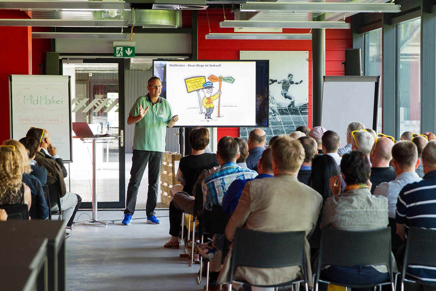 Ein Mann hält einen Vortrag vor einem sitzenden Publikum, das neben einem Bildschirm steht, auf dem ein Cartoon und zwei Flipcharts in einem hell erleuchteten Raum zu sehen sind.