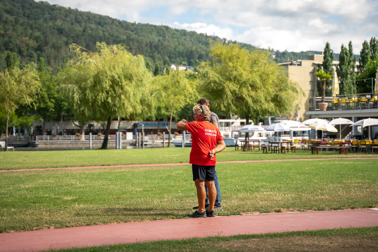Zwei Personen stehen auf einer Wiese in der Nähe einer Rennstrecke; eine trägt ein rotes Hemd mit weißer Schrift. Im Hintergrund sind Bäume, Boote und Sitzgelegenheiten im Freien zu sehen.