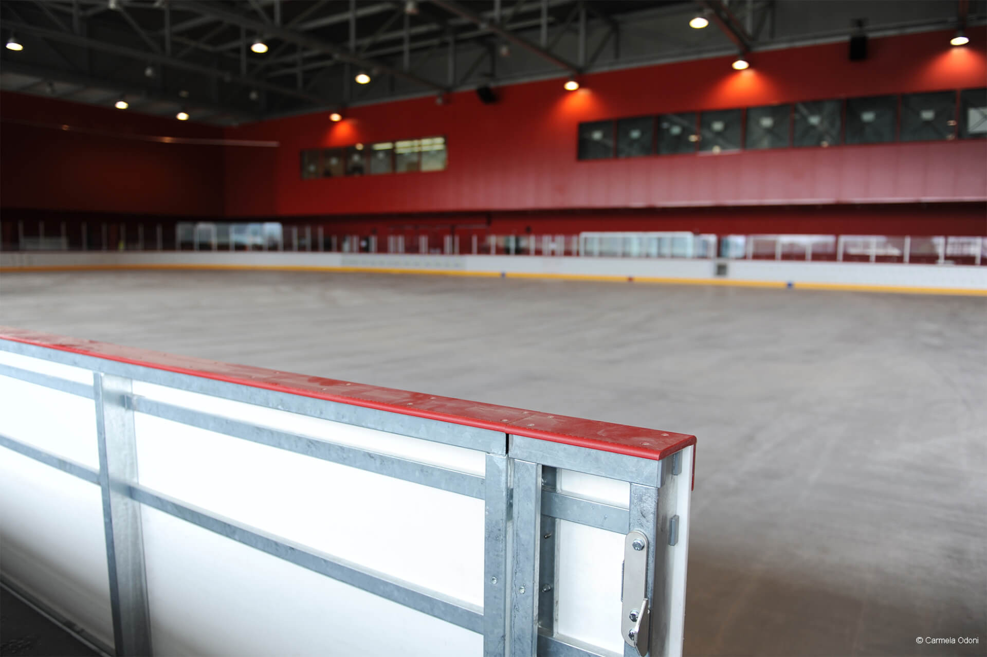 Eine überdachte Eisbahn mit roten Wänden, ohne Eis und Menschen, von hinten gesehen.