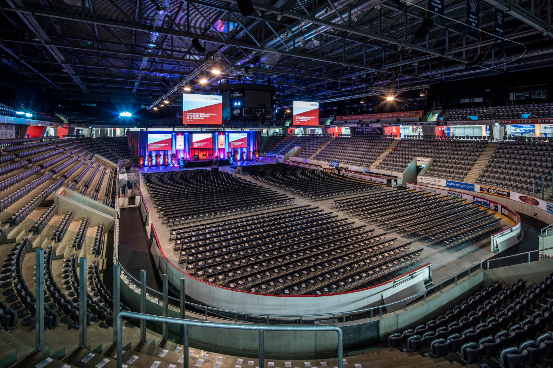 Eine leere Halle mit Reihen schwarzer Stühle vor einer Bühne mit großen Bildschirmen und Bannern bei heller Beleuchtung.