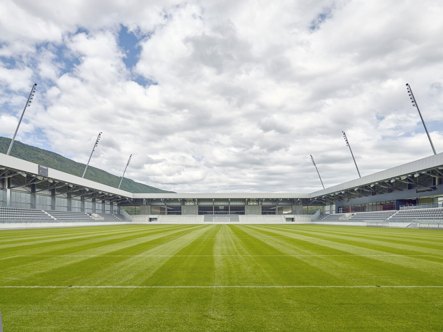 Leeres Fußballstadion mit grünem Rasen, umliegenden Tribünen und Oberlichtern unter einem teilweise bewölkten Himmel.