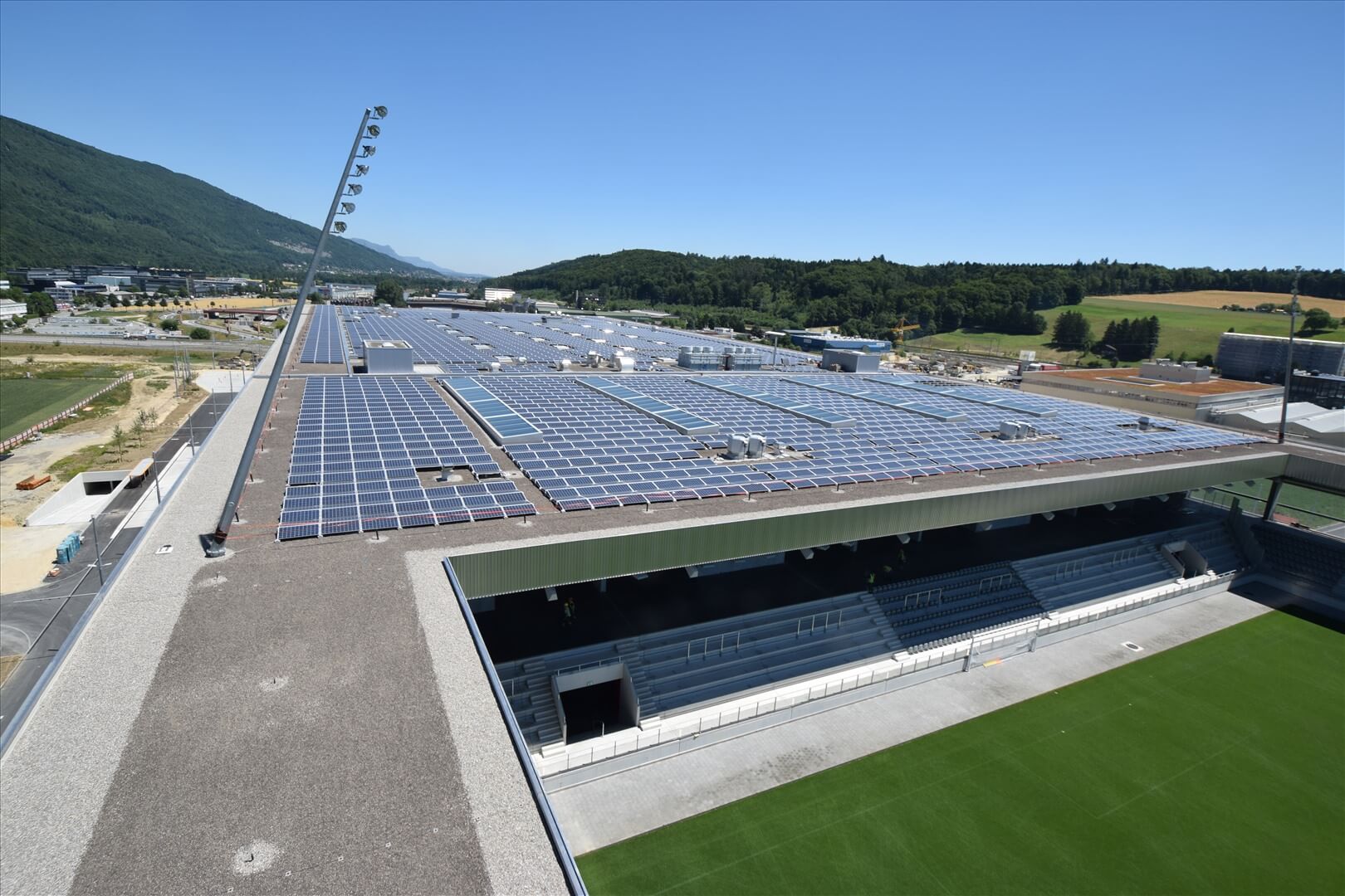Ein großes Sportstadion mit einem mit Sonnenkollektoren bedeckten Dach, angrenzend an grüne Felder und umgeben von Bäumen und Hügeln unter einem klaren blauen Himmel.