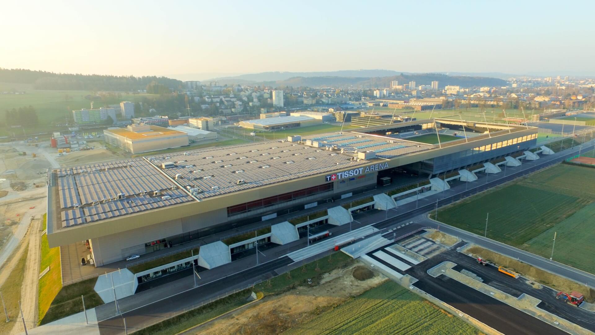 Luftaufnahme der Tissot Arena, eines großen modernen Sportkomplexes mit Sonnenkollektoren auf dem Dach, umgeben von Straßen, Grünflächen und städtischen Gebäuden im Hintergrund.