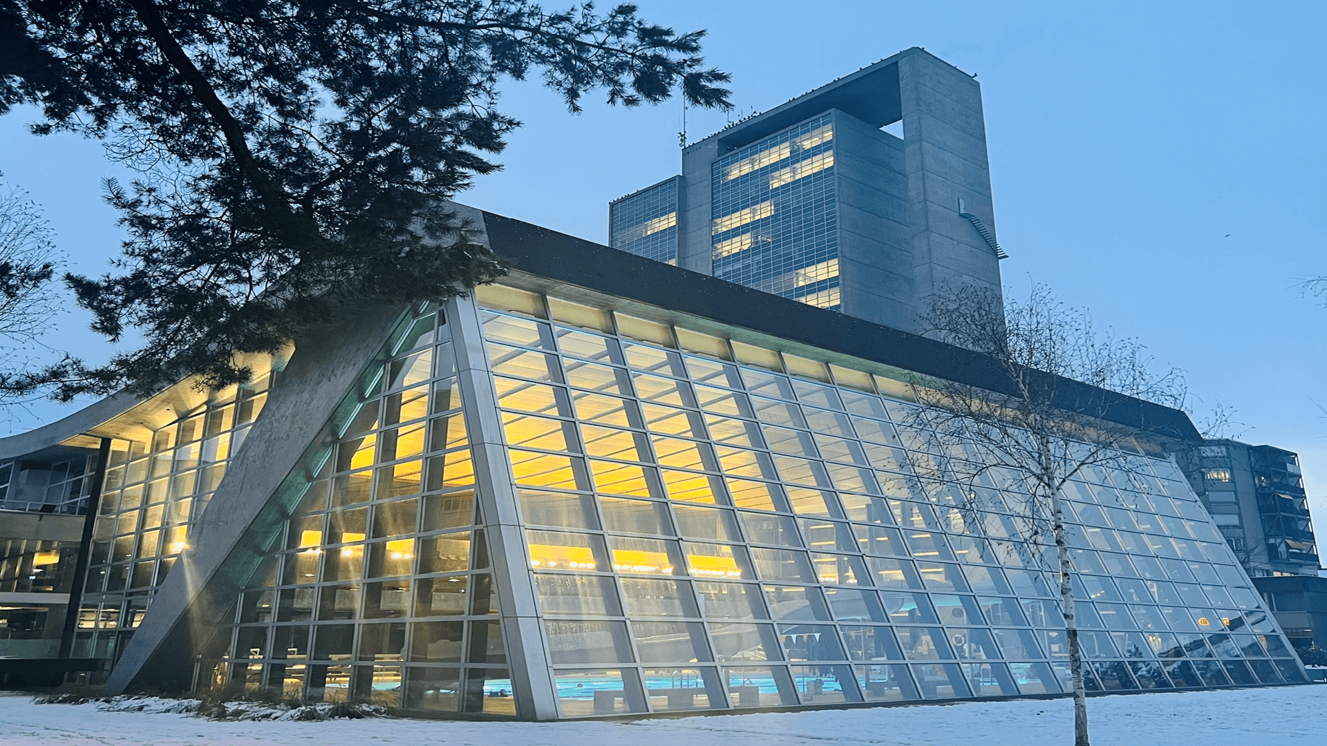 Ein modernes Gebäude mit einer großen Glasfassade wird in der Abenddämmerung von innen beleuchtet, umgeben von Schnee und ein paar Bäumen.