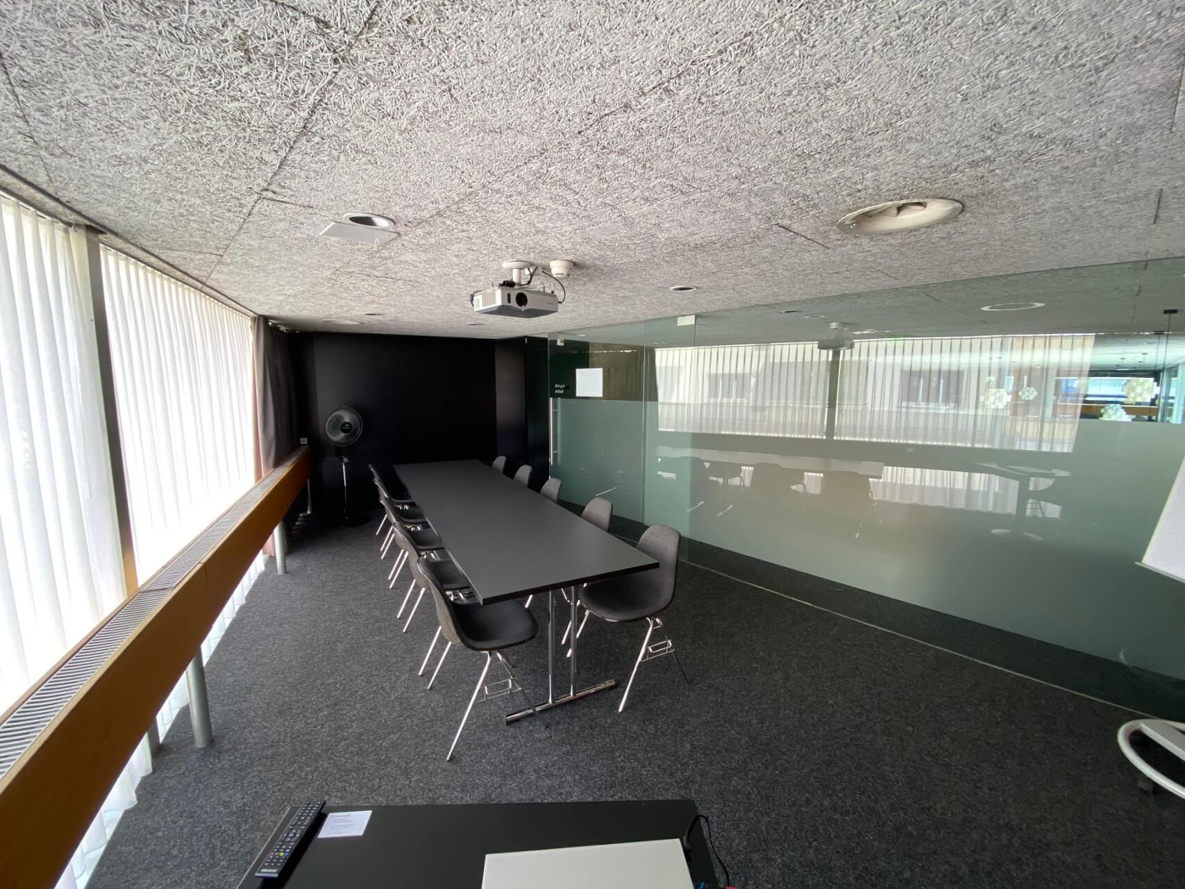 Ein moderner Konferenzraum mit einem langen schwarzen Tisch, mehreren Stühlen, einem Projektor an der Decke und großen Fenstern mit Jalousien, die natürliches Licht hereinlassen.