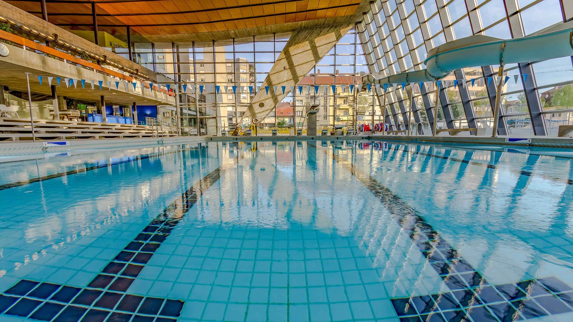 Hallenbad mit Bahnen, blauen und weißen Fahnen über dem Becken, großen Glasfenstern und einer Wasserrutsche auf der rechten Seite.