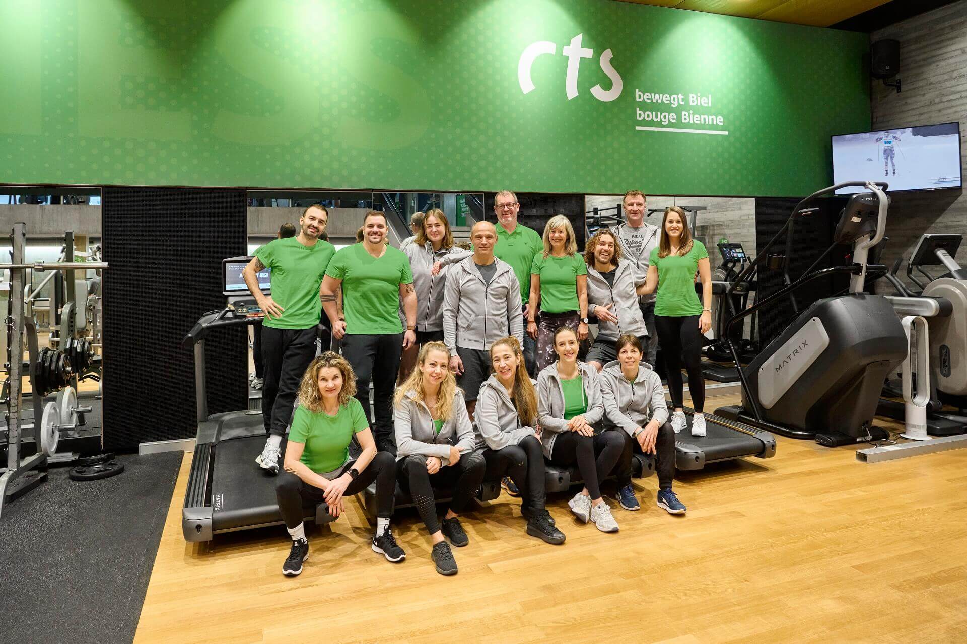 Eine Gruppe von Menschen in grüner und grauer Sportkleidung posiert für ein Foto in einem modernen Fitnessstudio. Sie stehen und sitzen vor Laufbändern und Trainingsgeräten.