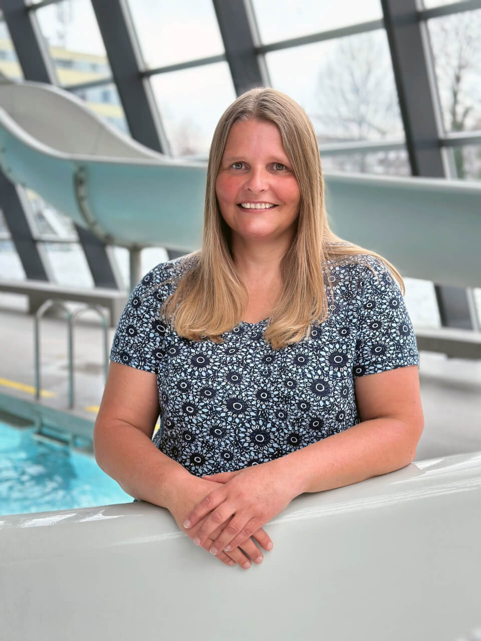 Eine Frau mit glattem blondem Haar und einem gemusterten Hemd steht lächelnd in der Nähe eines Hallenbades mit einer Rutsche im Hintergrund.