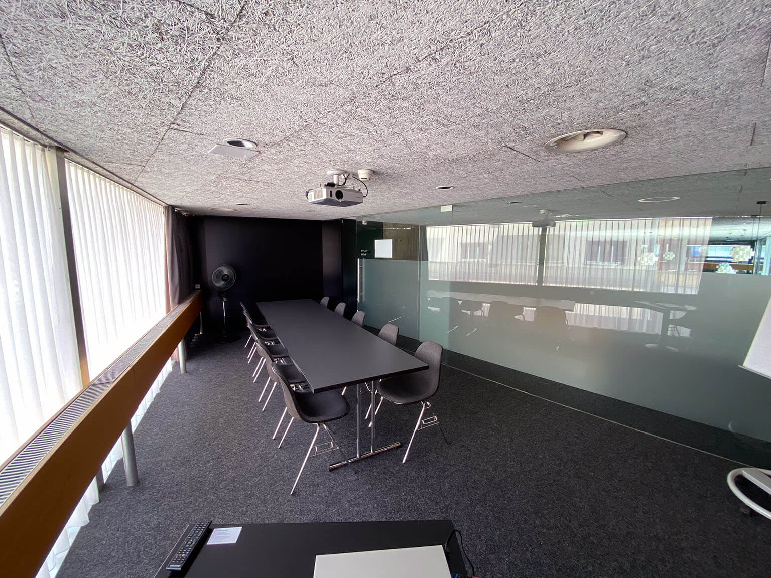 Ein moderner Konferenzraum mit einem langen schwarzen Tisch, mehreren Stühlen, einem Projektor an der Decke und großen Fenstern mit Jalousien, die natürliches Licht hereinlassen.