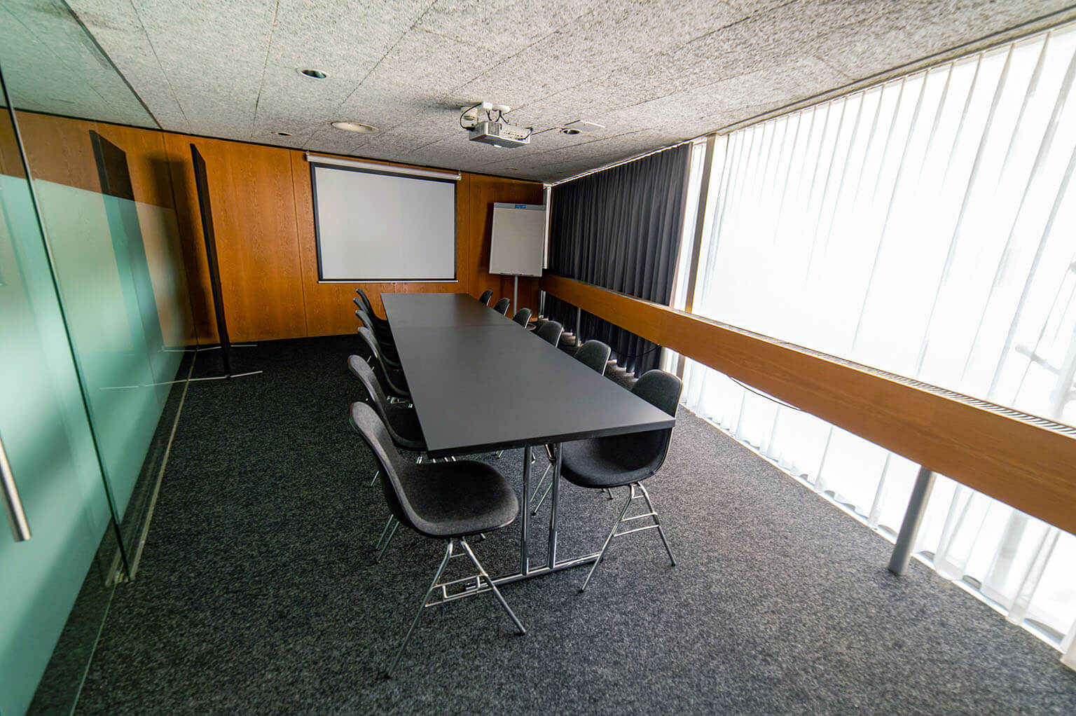 Moderner Konferenzraum mit einem langen schwarzen Tisch, zehn Stühlen, einem Projektor, einer Projektionsfläche, einem Flipchart, Glaswänden und großen Fenstern mit vertikalen Jalousien.