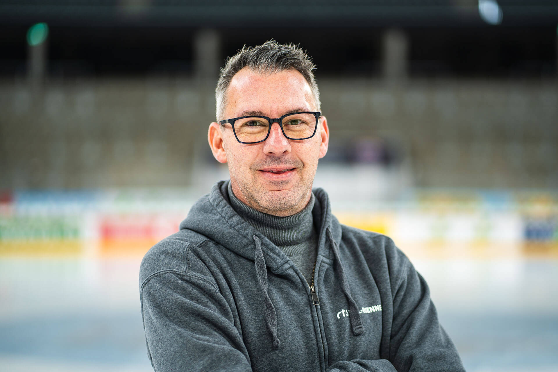 Ein Mann mittleren Alters mit Brille und kurzen Haaren steht in einem Innenraum, trägt einen grauen Kapuzenpulli und einen Rollkragenpullover - und fängt einen Moment aus dem Alltag vom Leiter Eismeister in der Tissot Arena ein, mit unscharfen Stadionplätzen im Hintergrund.