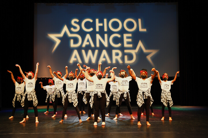 Eine Gruppe von Tänzerinnen und Tänzern in passenden Outfits tritt auf der Bühne vor einem großen, auf den Hintergrund projizierten School Dance Award-Schild auf.