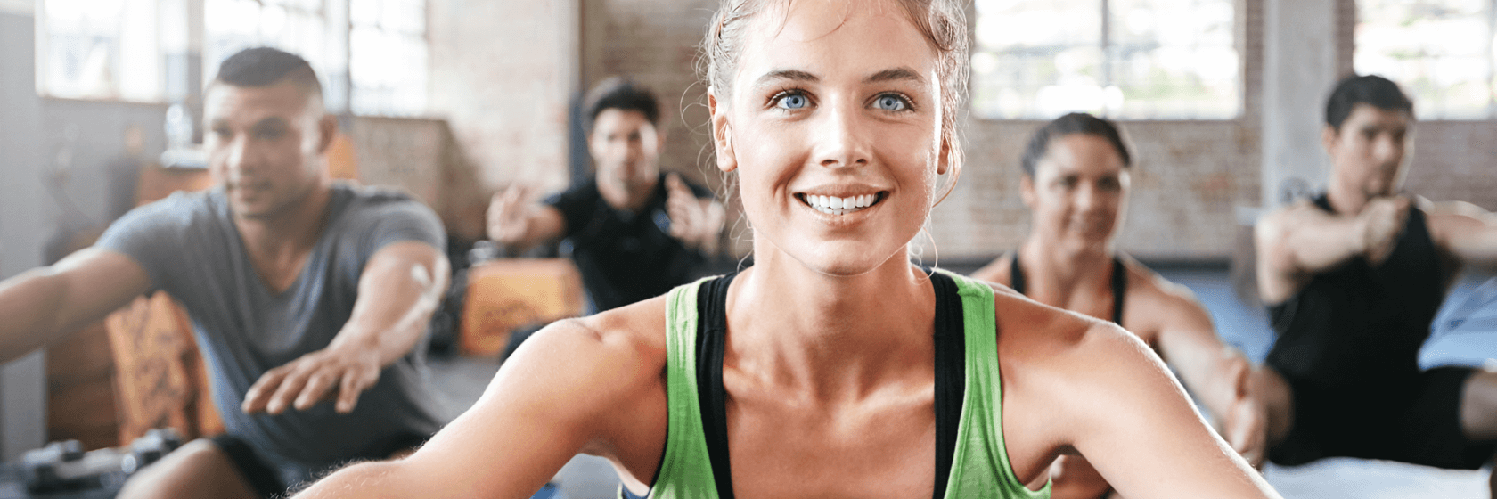 Eine Gruppe von Menschen, die in einer Halle trainieren, mit einer Frau in einem grünen Tank Top, die vorne lächelt, und anderen, die im Hintergrund Kniebeugen machen.