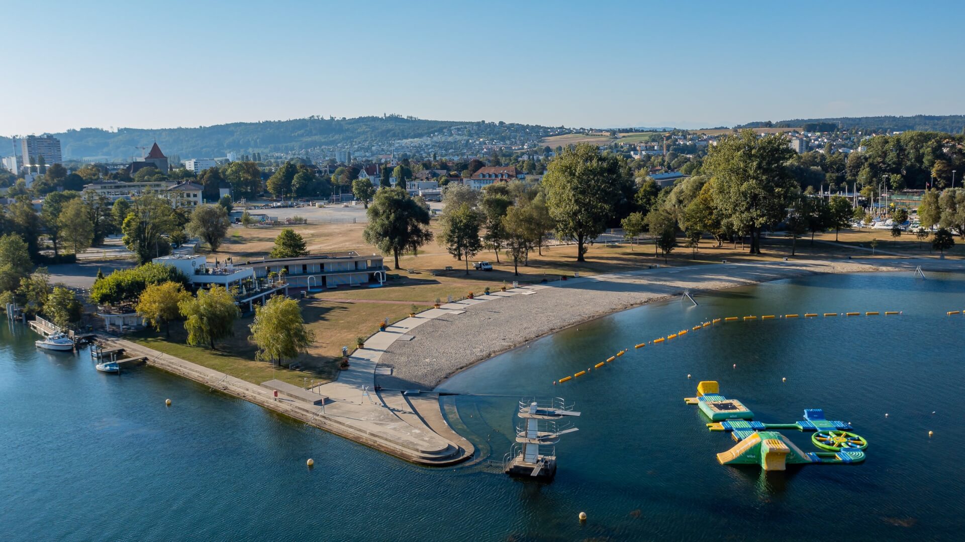Ein Park am See mit Sandstrand, Bäumen, einem Spielplatz am Wasser und nahe gelegenen Gebäuden unter freiem Himmel.