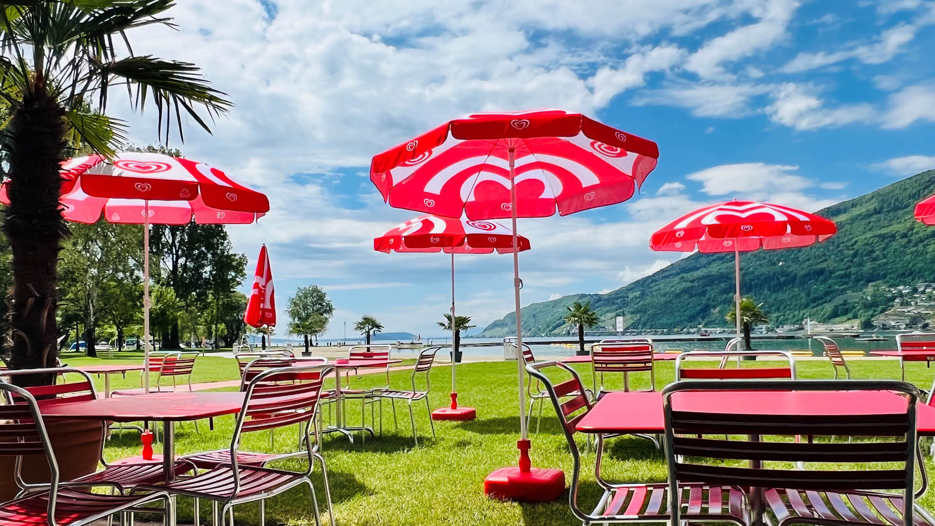 Außencafé mit roten Tischen, Stühlen und Sonnenschirmen auf einer Wiese am See, mit Bergen und blauem Himmel im Hintergrund.