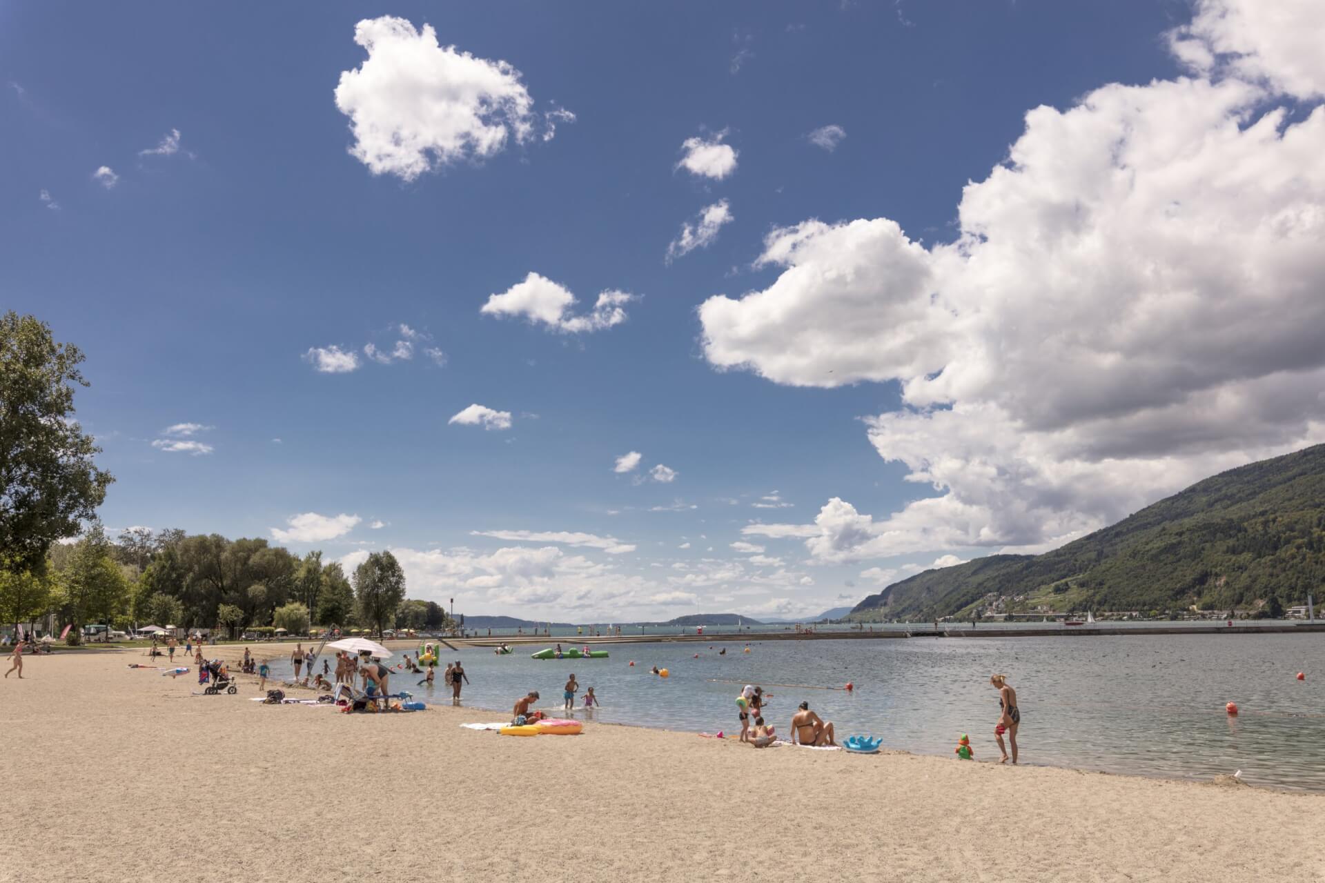 An einem sonnigen Tag mit teilweise bewölktem Himmel entspannen und spielen Menschen an einem Sandstrand neben einem See mit fernen Hügeln.