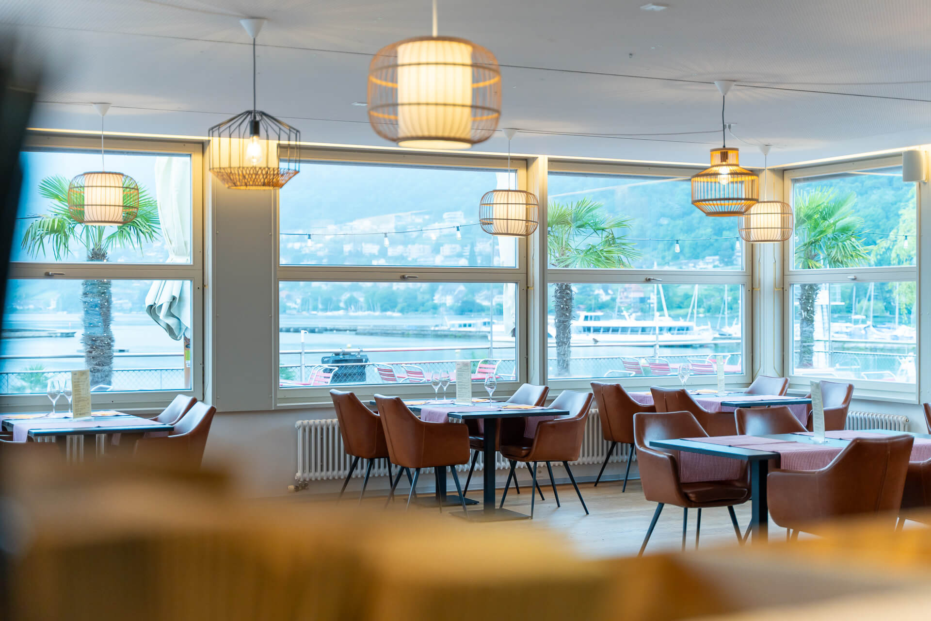 Modernes Restaurant mit braunen Stühlen, quadratischen Tischen und großen Fenstern, die den Blick auf Palmen, den Yachthafen und die Hügel im Hintergrund freigeben.