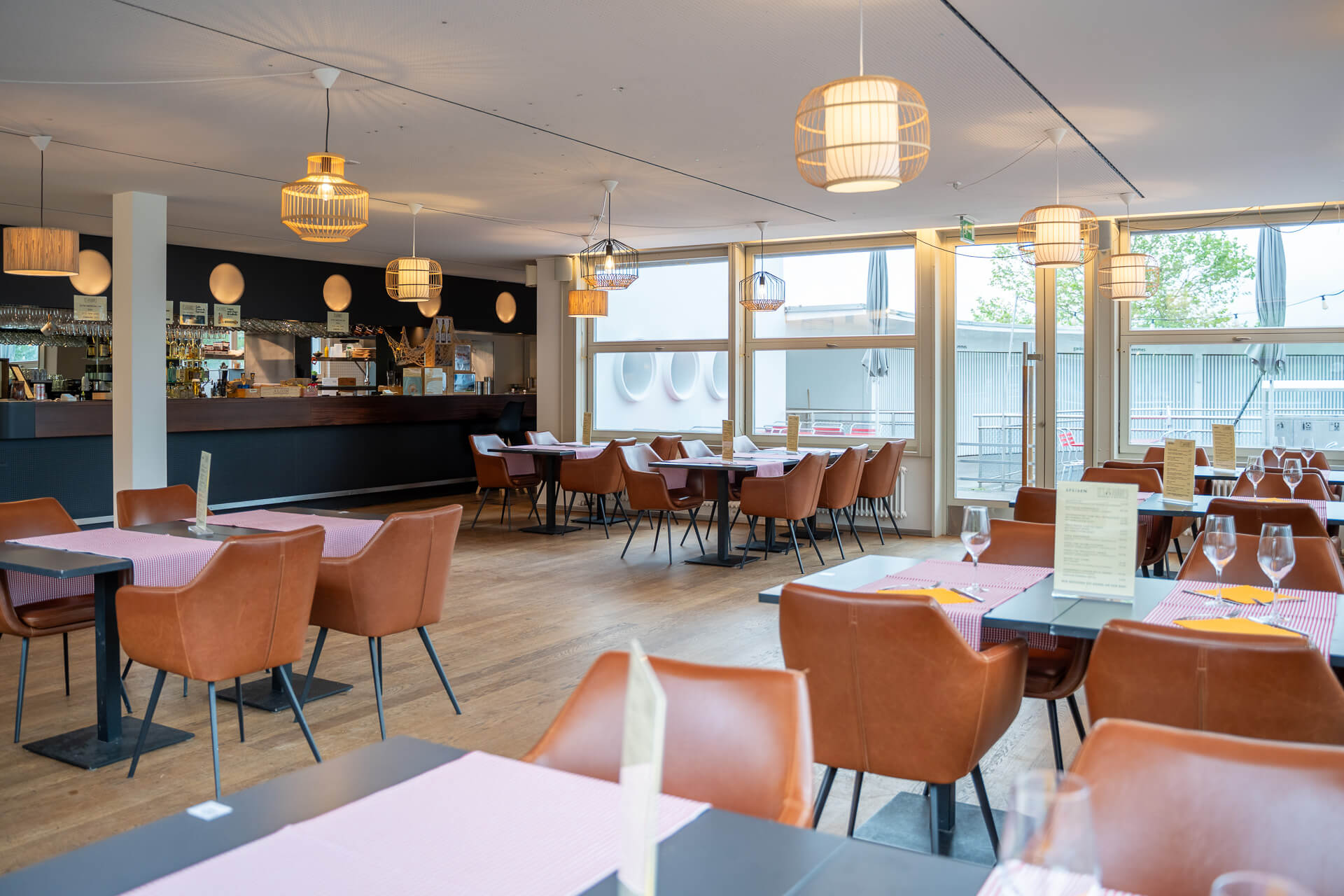 Modernes Restaurantinterieur mit braunen Lederstühlen, gedeckten Tischen mit Gläsern und Servietten, großen Fenstern, Holzboden und Hängeleuchten.