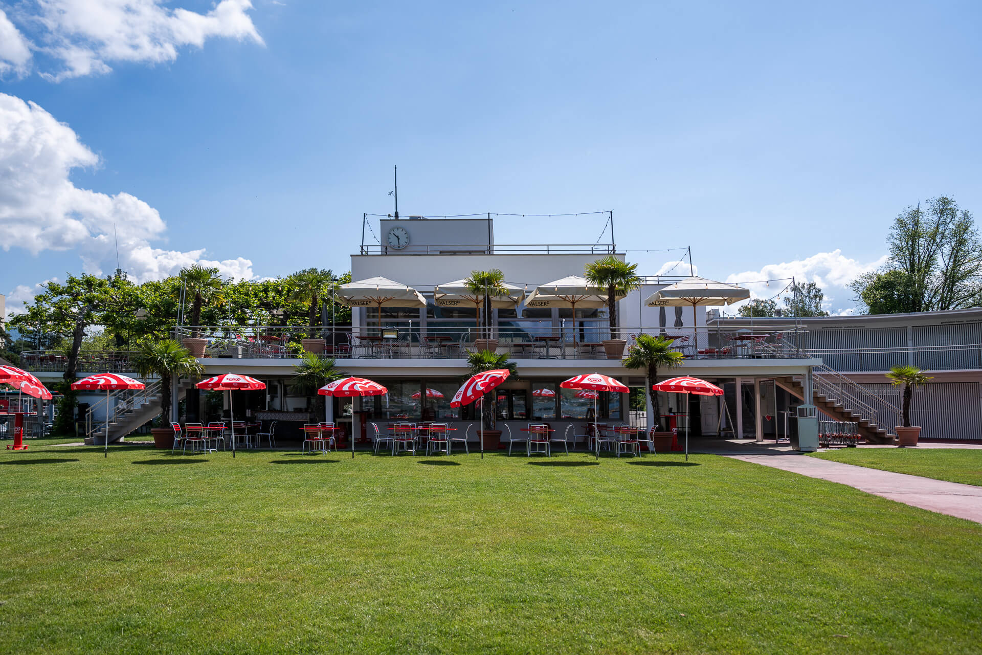 Modernes zweistöckiges Gebäude mit Sitzgelegenheiten im Freien und roten Sonnenschirmen auf einer grünen Wiese unter einem blauen Himmel mit vereinzelten Wolken.