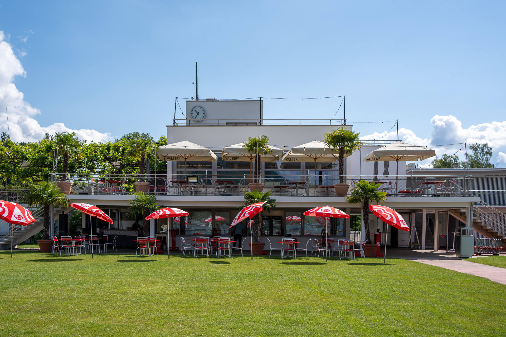 Modernes zweistöckiges Gebäude mit Sitzgelegenheiten im Freien, roten und weißen Sonnenschirmen, Palmen und einer Uhr auf dem Dach, auf einer Wiese unter blauem Himmel.