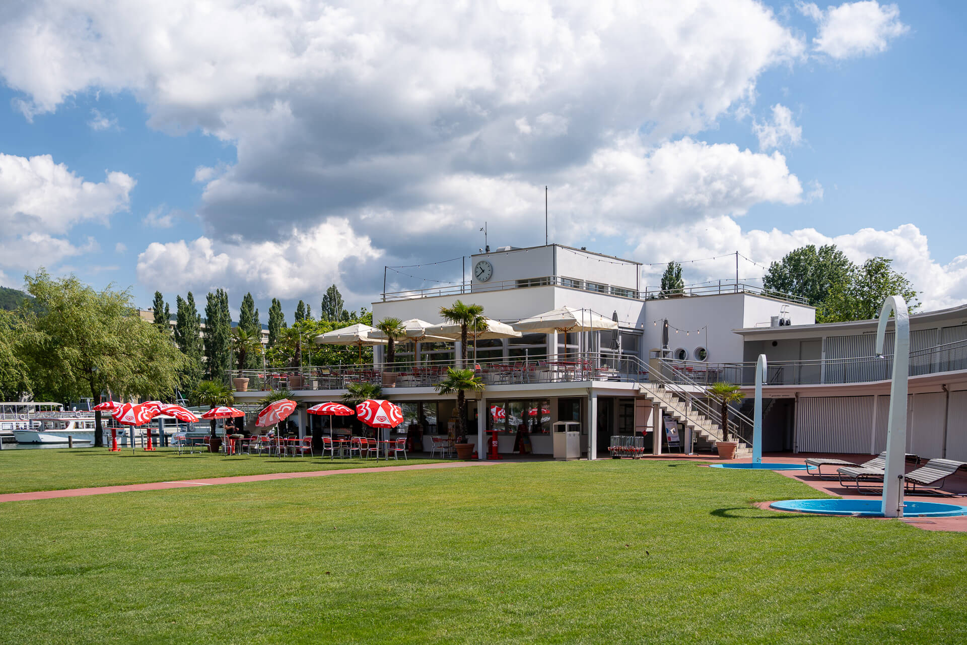 Ein zweistöckiges modernes Gebäude mit Sitzgelegenheiten im Freien, roten Sonnenschirmen und einer großen Rasenfläche davor, unter einem teilweise bewölkten Himmel.
