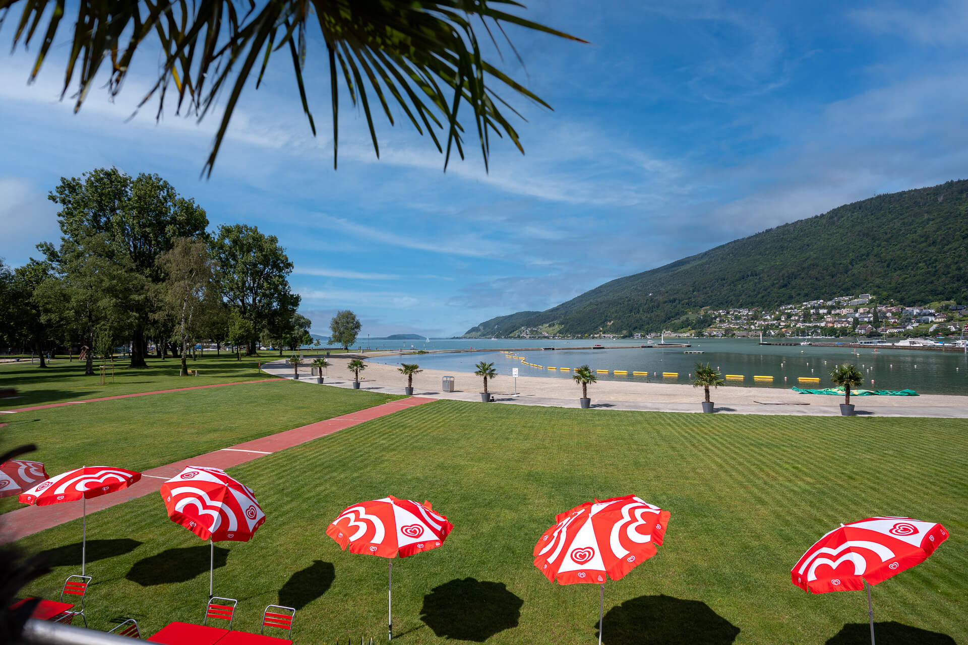 Ein grasbewachsener Park mit roten und weißen Sonnenschirmen überblickt die Uferpromenade mit den Bergen und der Stadt auf der anderen Seite der Bucht unter einem teilweise bewölkten Himmel.
